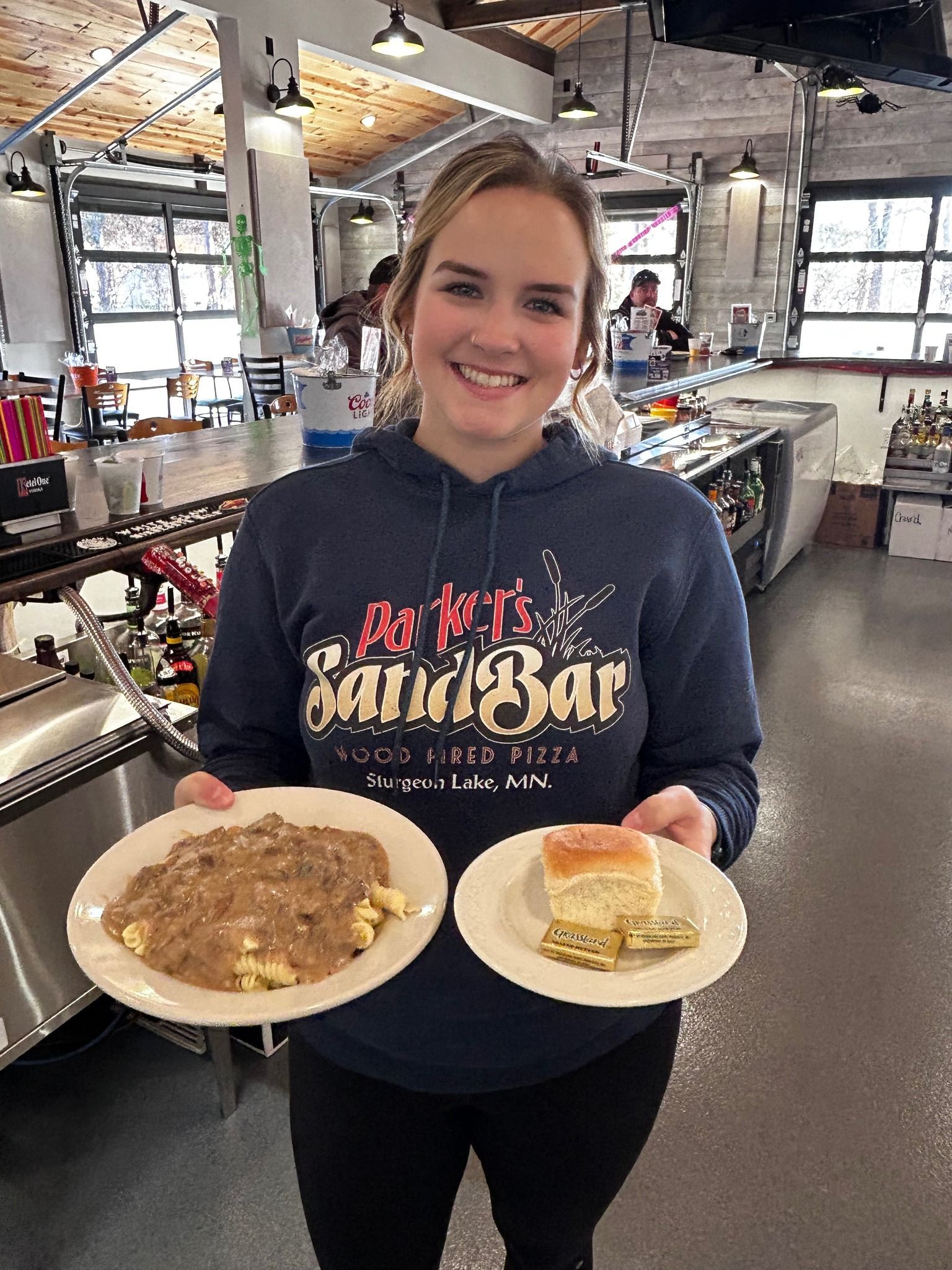 Smiling person in a navy hoodie holding two plates of food in a casual restaurant kitchen