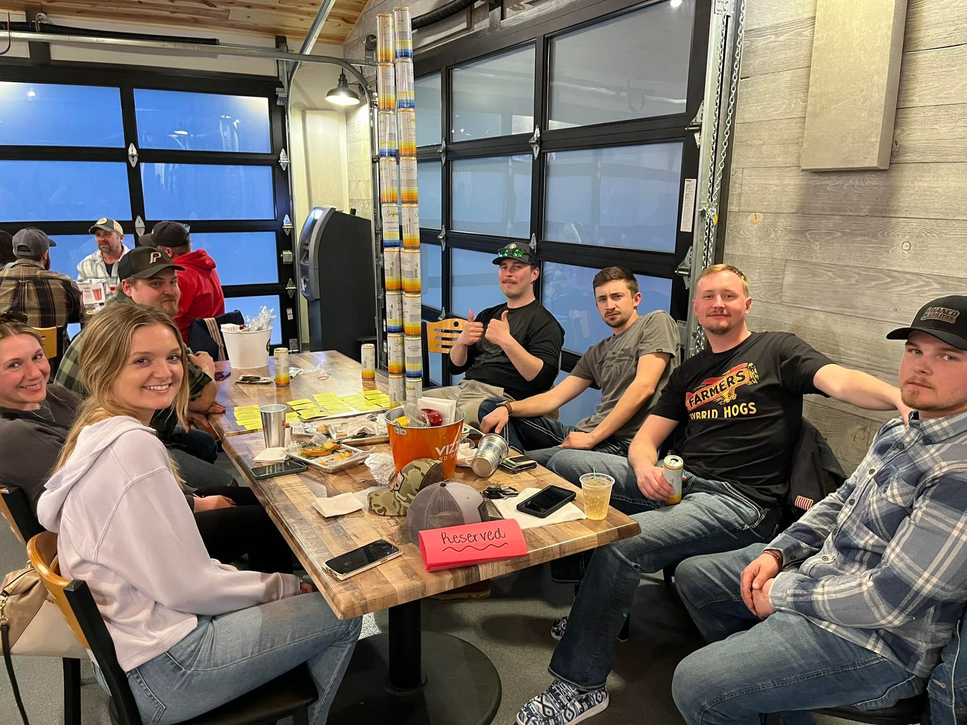 Group of people seated around a picnic table in a garage, smiling and raising drinks during a casual gathering