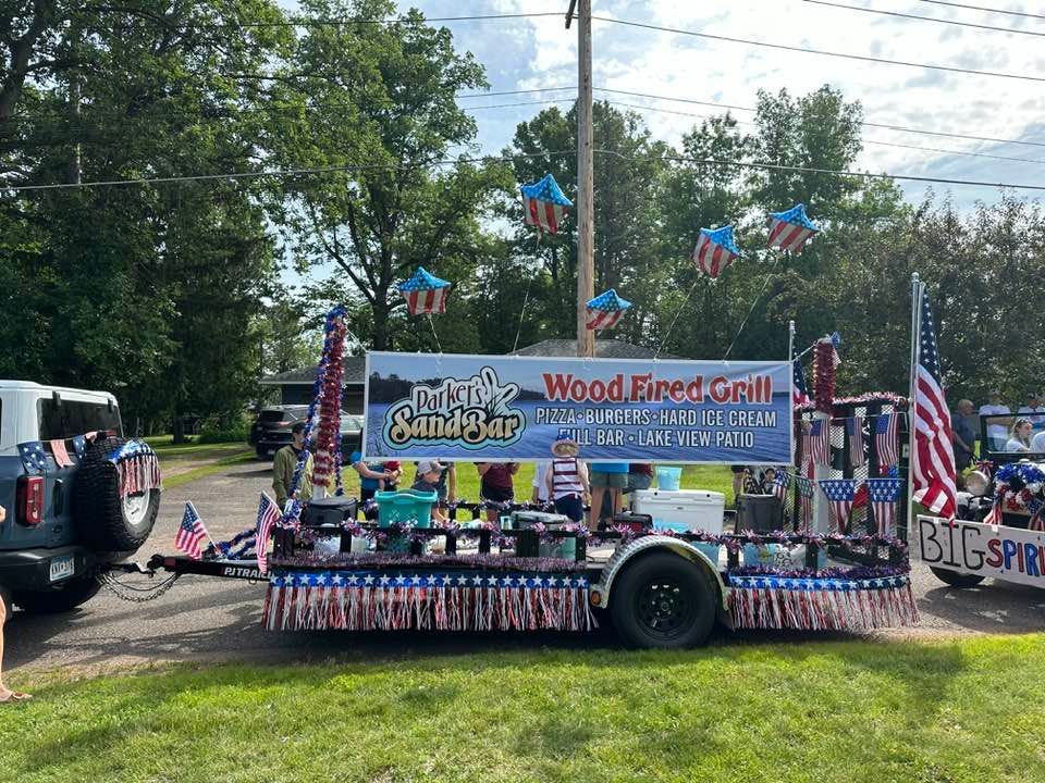 Decorated parade float with patriotic bunting, flags, and people aboard, parked by a roadside under trees.