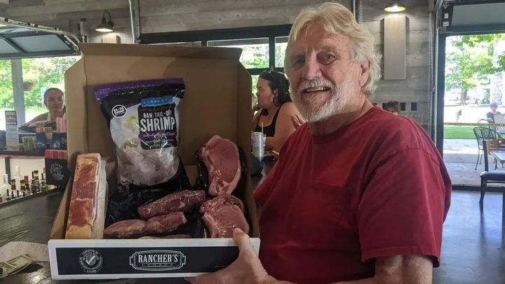 Smiling man in a red shirt holding a display of smoked meats at an indoor market booth