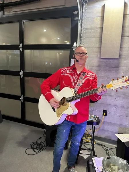 Man in a red shirt playing white acoustic guitar indoors near a wall of garage doors