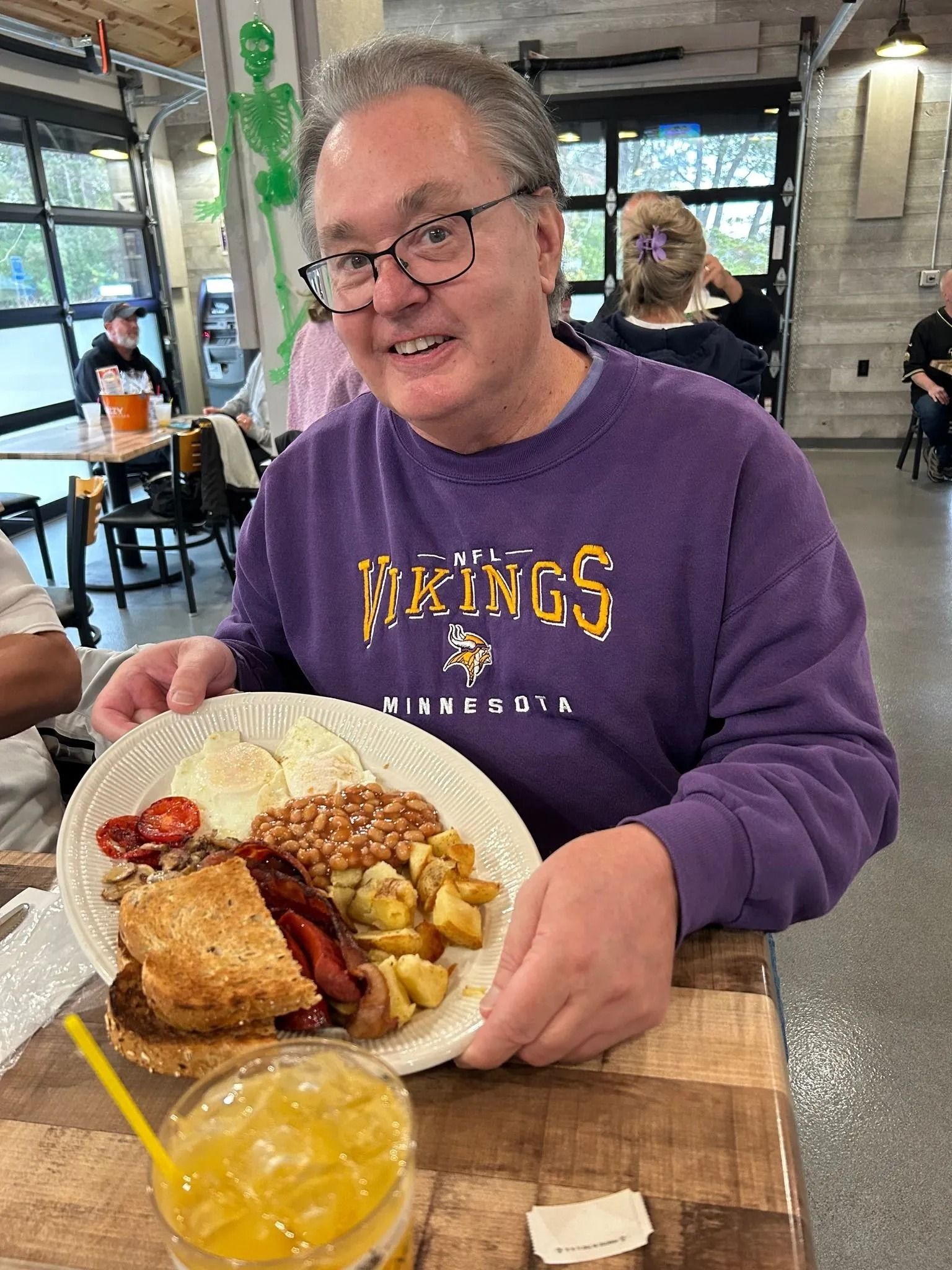 Smiling person in a purple Vikings shirt holding a plate of breakfast at a restaurant table