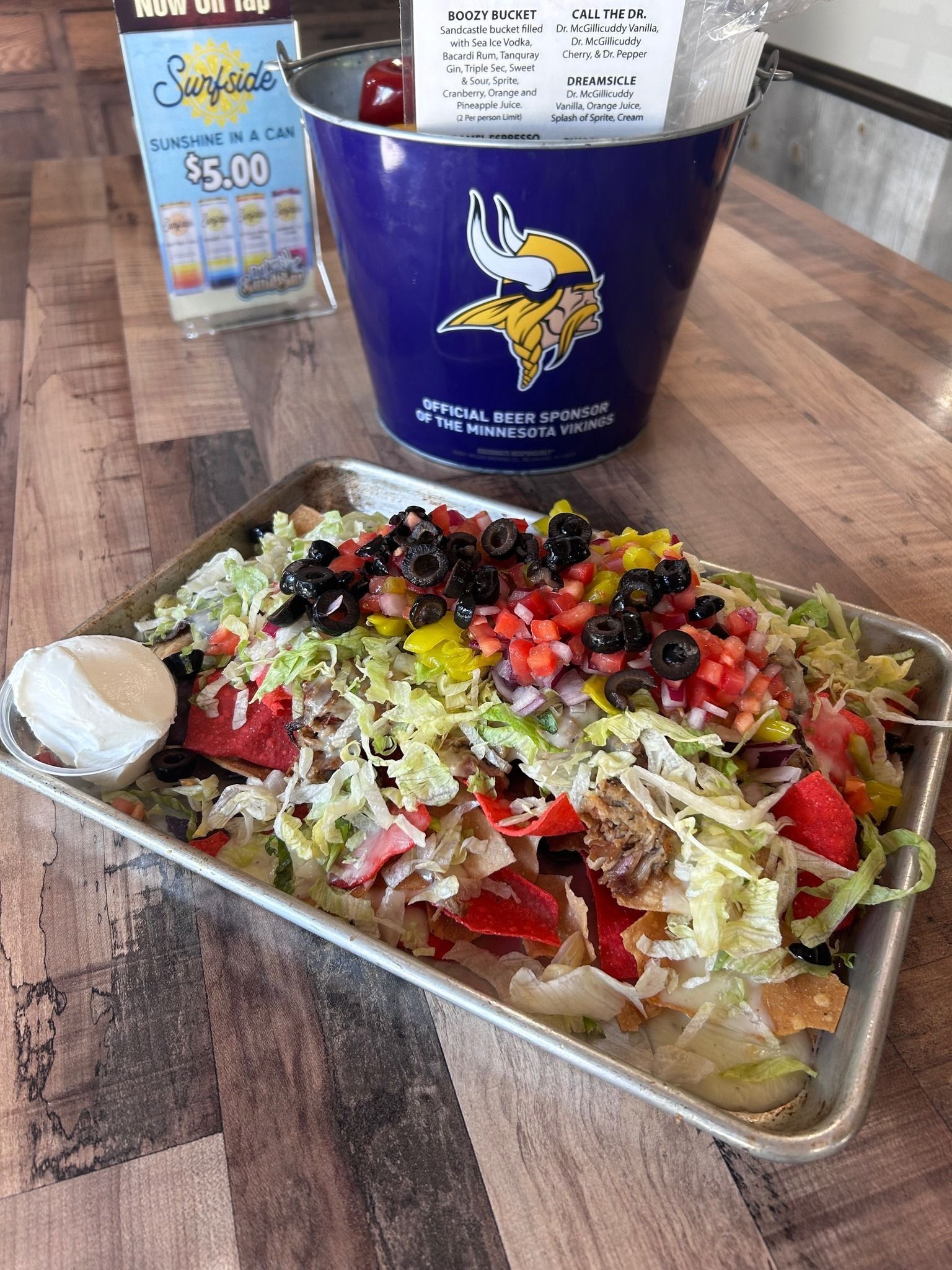 Loaded taco salad in a takeout tray with sour cream, beside a blue Minnesota Vikings cup on a table