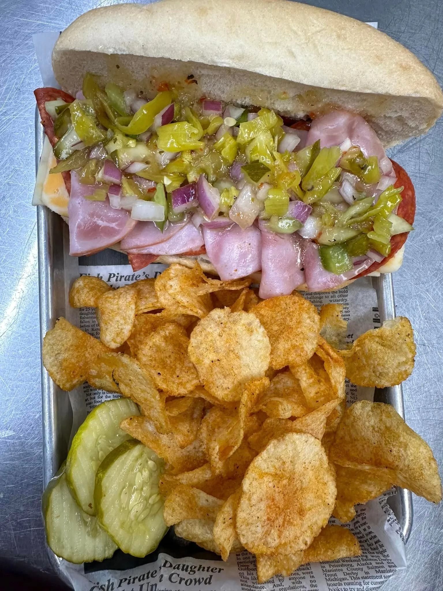 Sub sandwich with onions and peppers, served with seasoned potato chips and pickles on a tray.