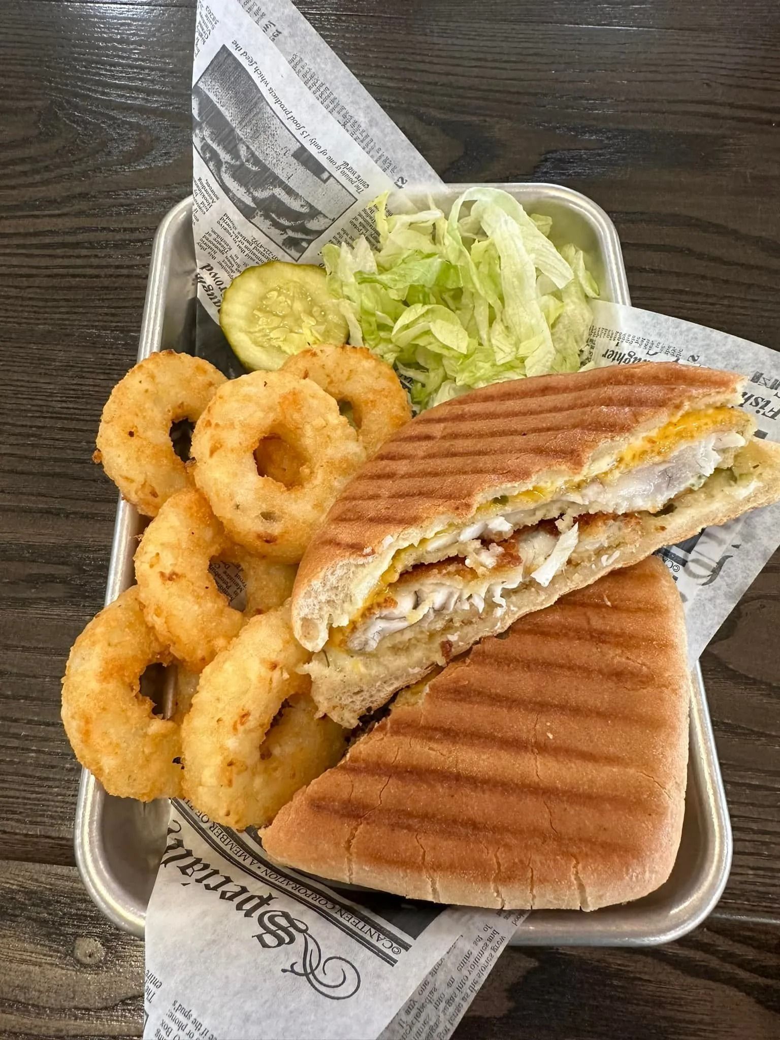 Grilled sandwich with onion rings, lettuce, pickle, and chips in a takeout tray