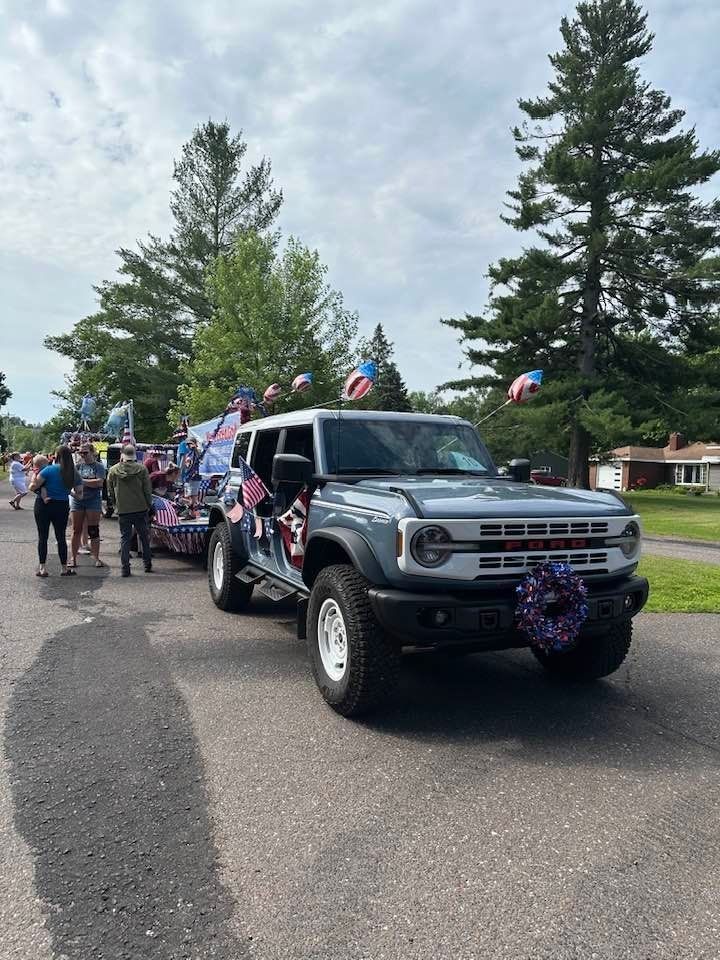 Decorated black SUV in a parade, with flags and people walking beside it on a tree-lined road