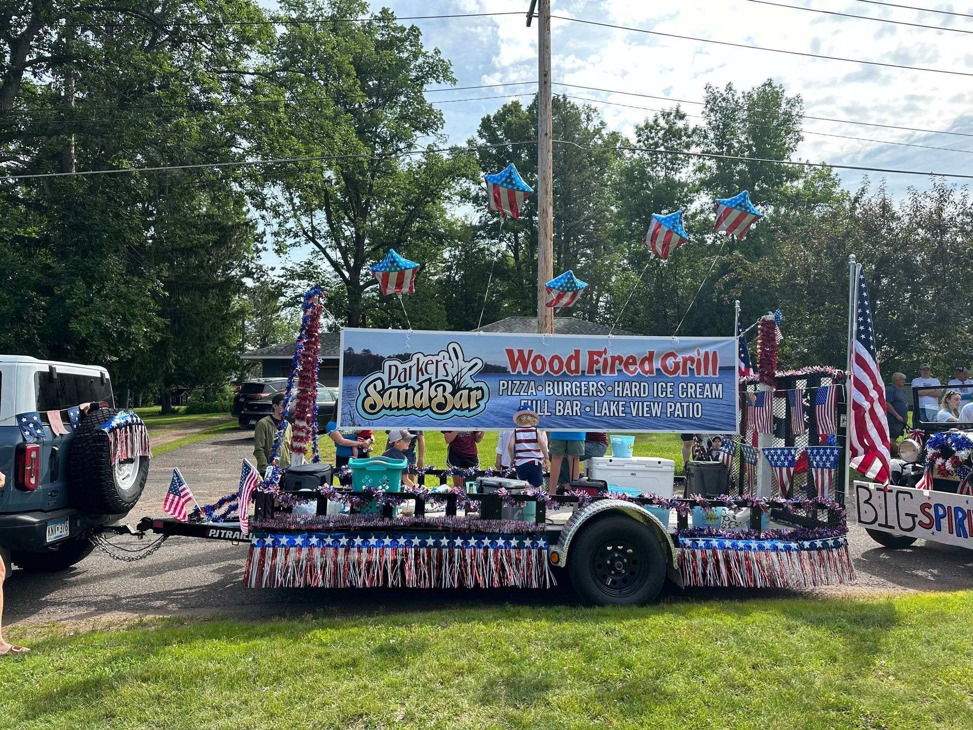 Decorated parade float with patriotic signs, flags, and people seated inside on a sunny street.