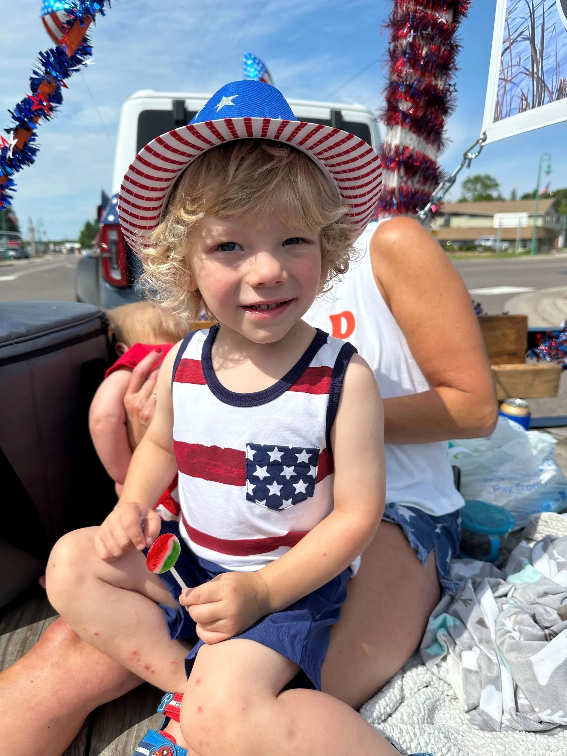 Child in patriotic outfit and sunhat sitting on an adult’s lap outdoors near water and boats.