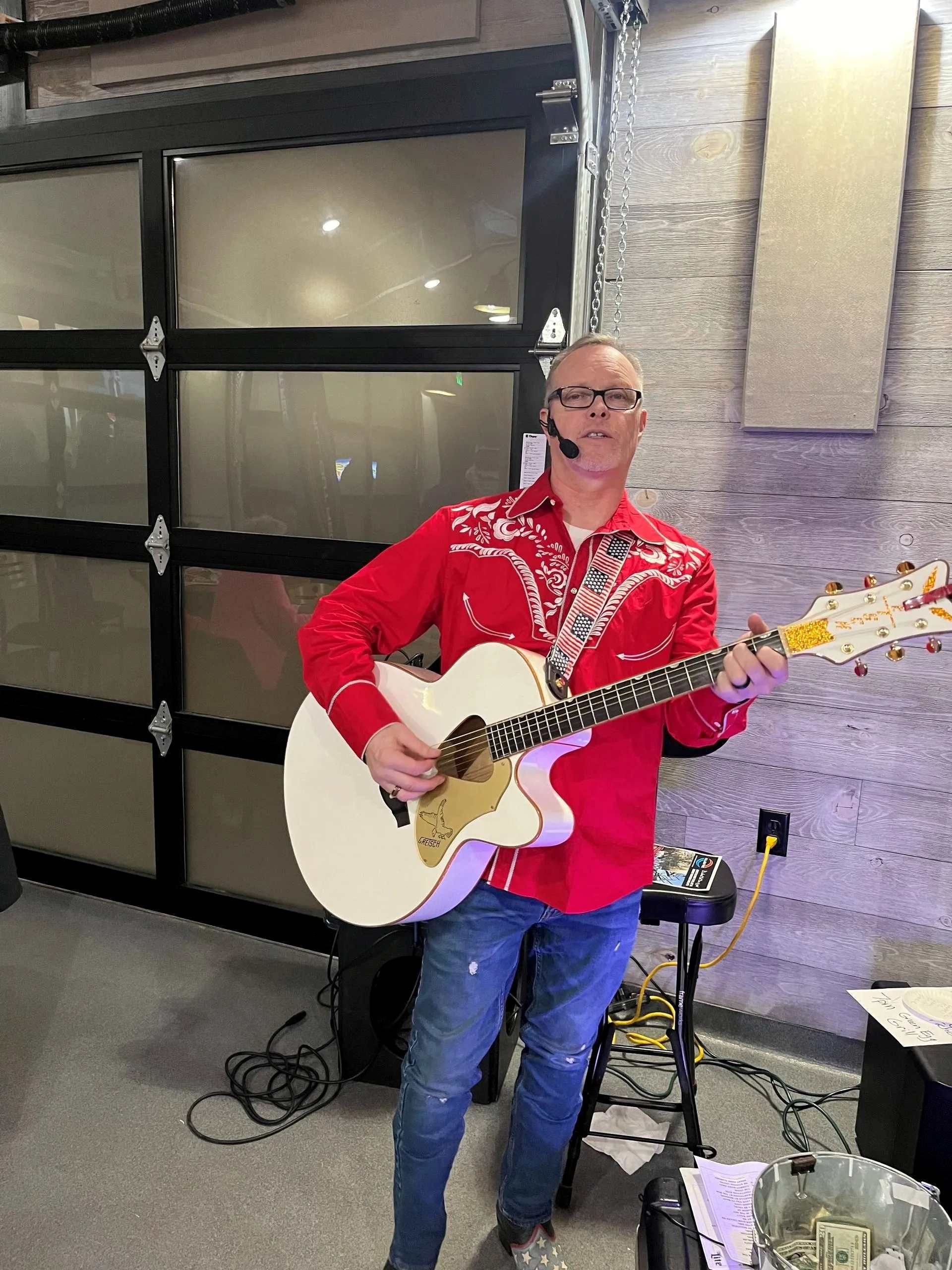 Man in a red shirt playing white acoustic guitar indoors beside a garage door