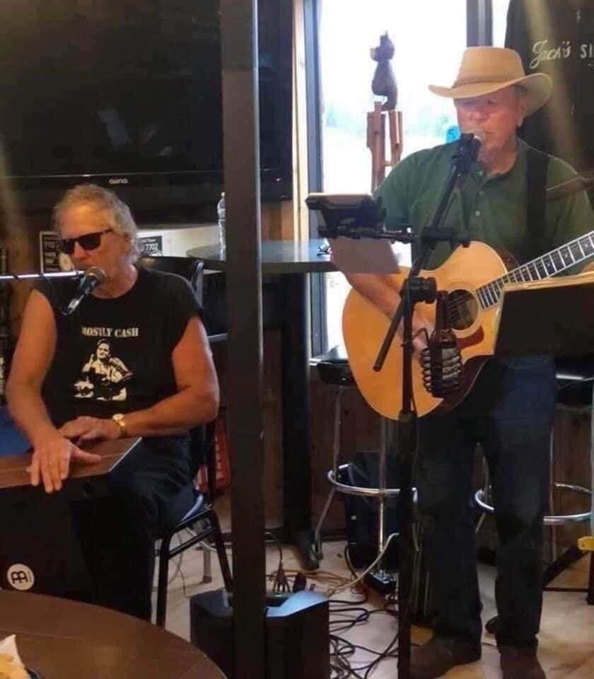 Two musicians performing indoors, one seated at a keyboard and one standing with an acoustic guitar.