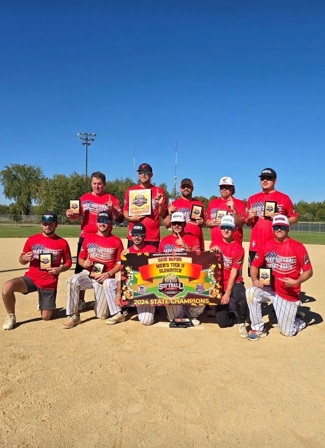Youth baseball team in red shirts posing with a championship banner on a sunny field.