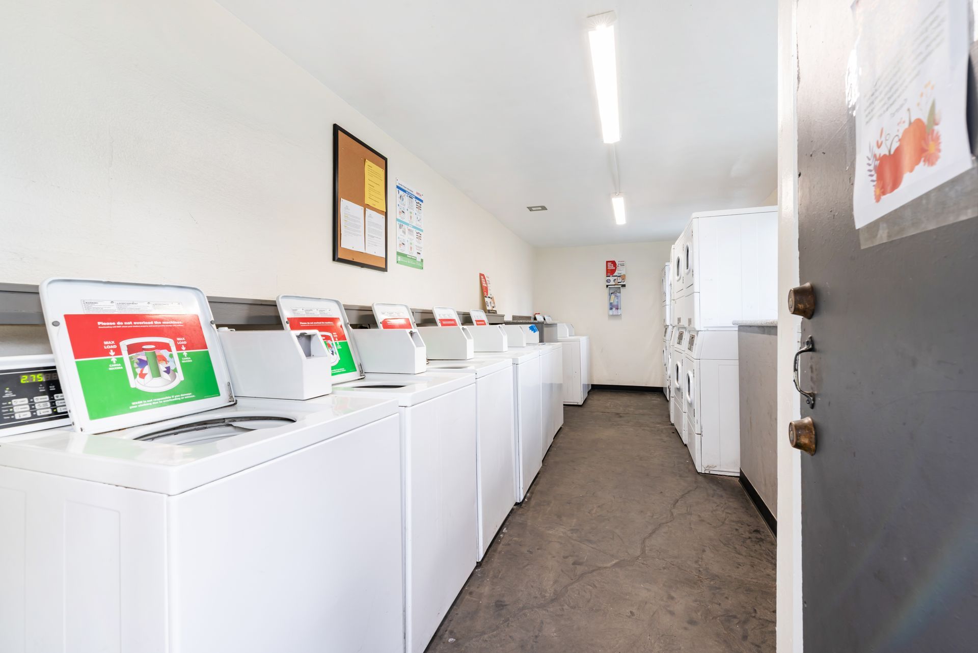 Row of white washing machines in a laundromat, with posters and a door on the right.