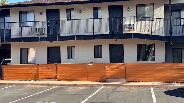 Apartment building with two floors, black doors, and a brown fence in front, viewed from a parking lot.
