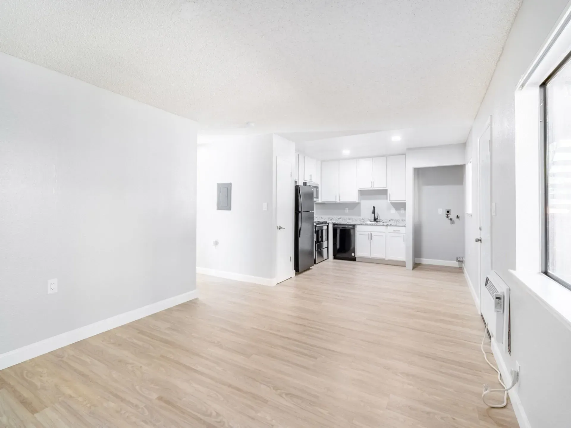 Empty, open-plan apartment interior with light wood flooring, white walls, and a small kitchen visible in the background.