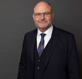 Professionelt portretfoto af en person med briller, et marineblåt jakkesæt og et mønstret slips mod en grå væg.