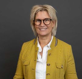 En smilende person med blond hår og briller, iført en hvid skjorte og en gulmønstret blazer mod en mørk baggrund.
