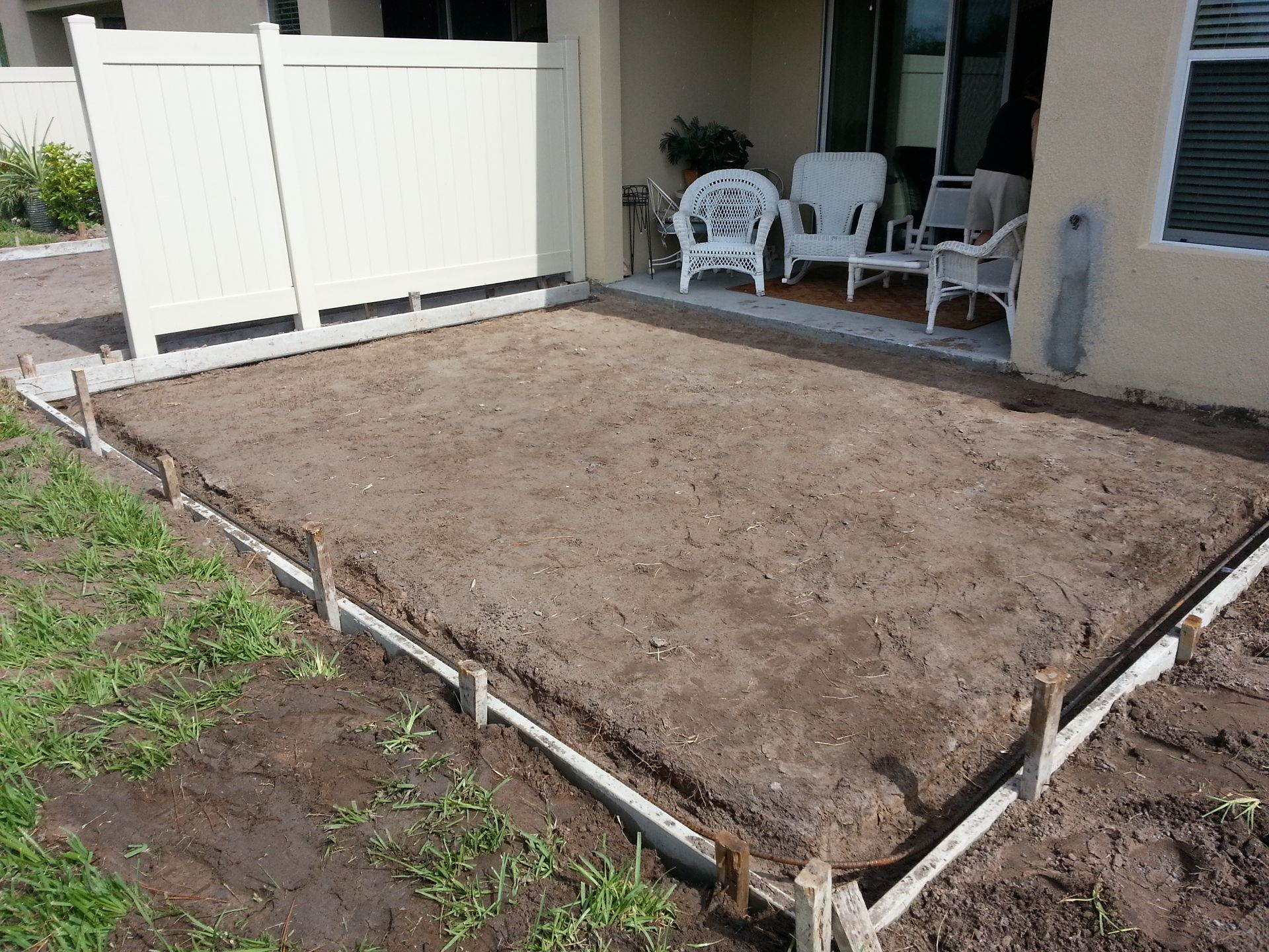 Freshly leveled dirt patio area beside a house, bordered by white fencing and patio chairs in the background