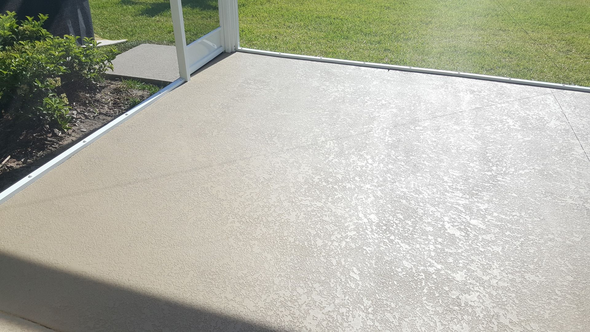 Concrete patio with sunlight, bordered by grass and garden plants near a glass railing.