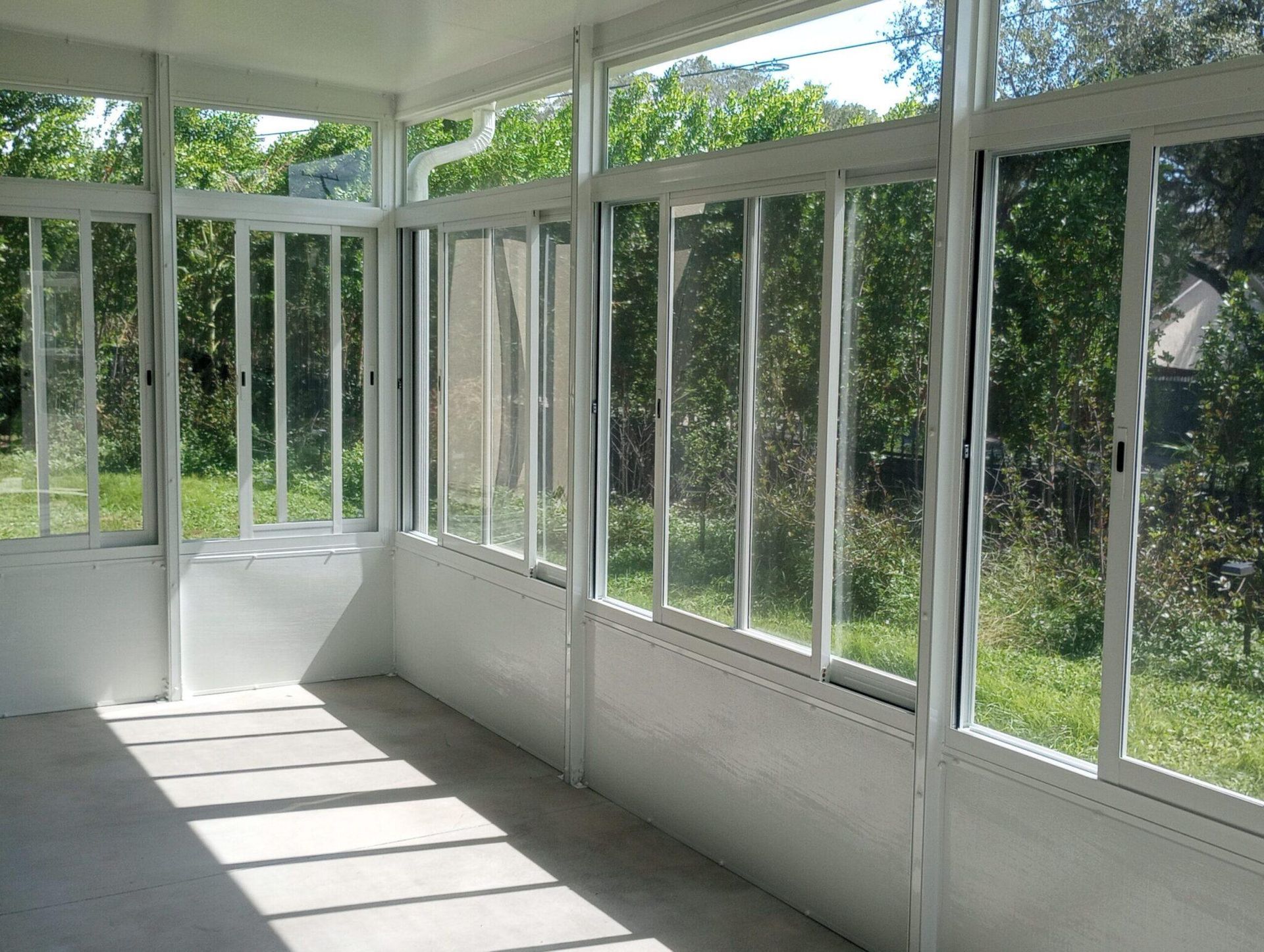 Sunlit enclosed porch with white-framed windows and doors overlooking green trees and grass
