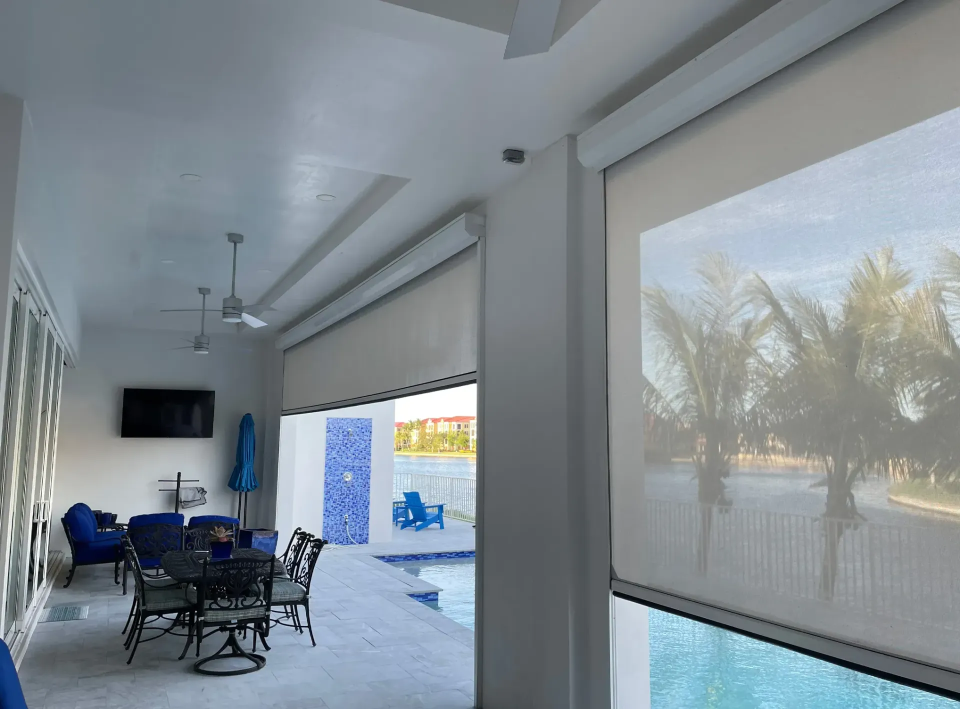 Covered patio with blue chairs, wall-mounted TV, and large windows overlooking palm trees and a pool