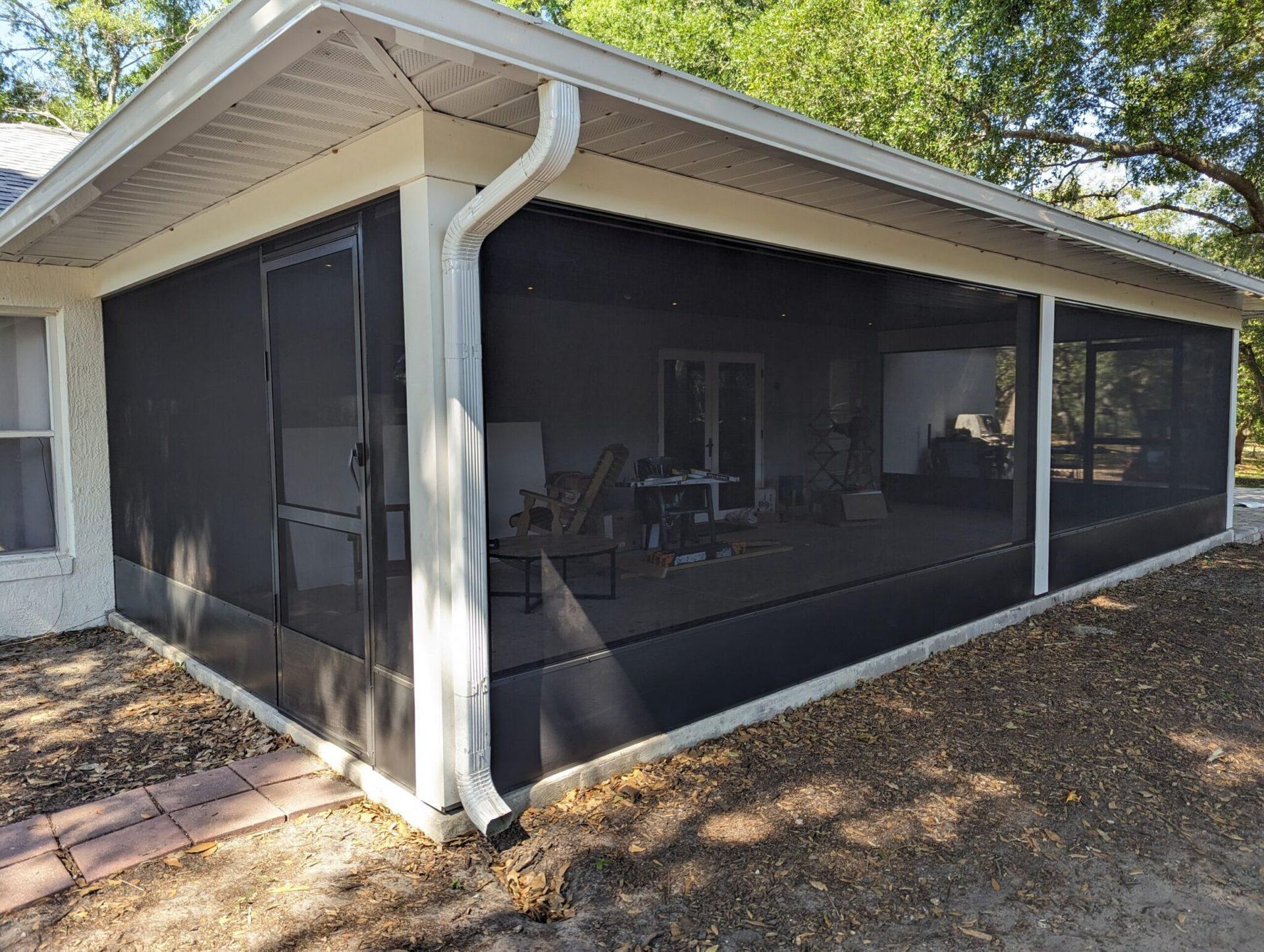 Screened porch with black mesh walls, white trim, and patio furniture beside a brick walkway under trees