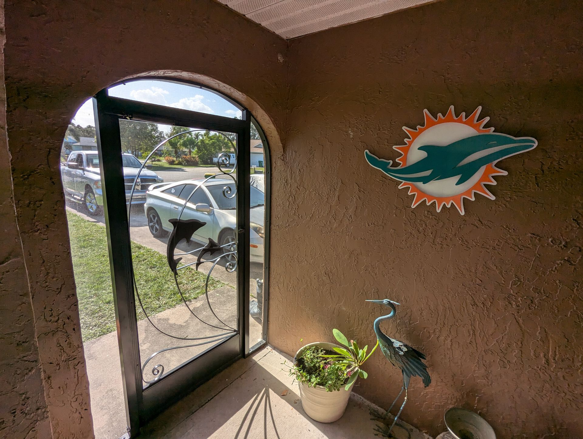 Front porch with open door, potted plant, metal flamingo decor, and a Miami Dolphins wall sign.