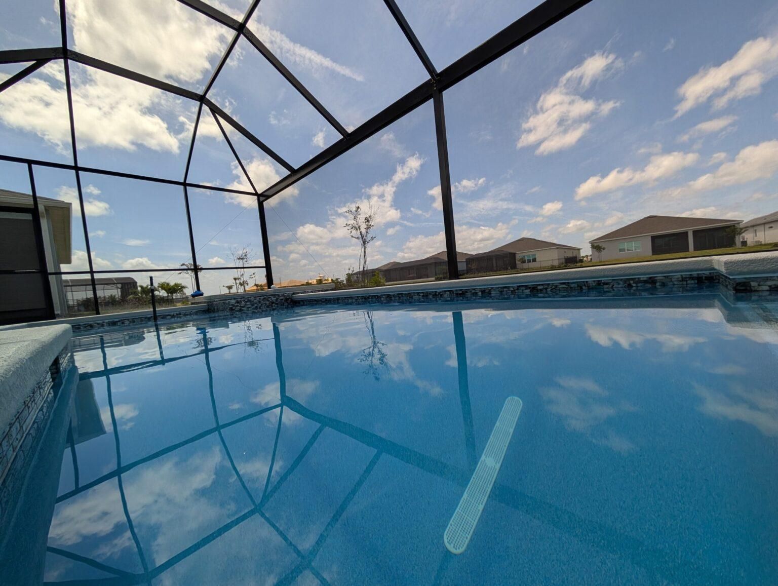 Screened-in backyard pool reflecting a blue sky with clouds and nearby houses.
