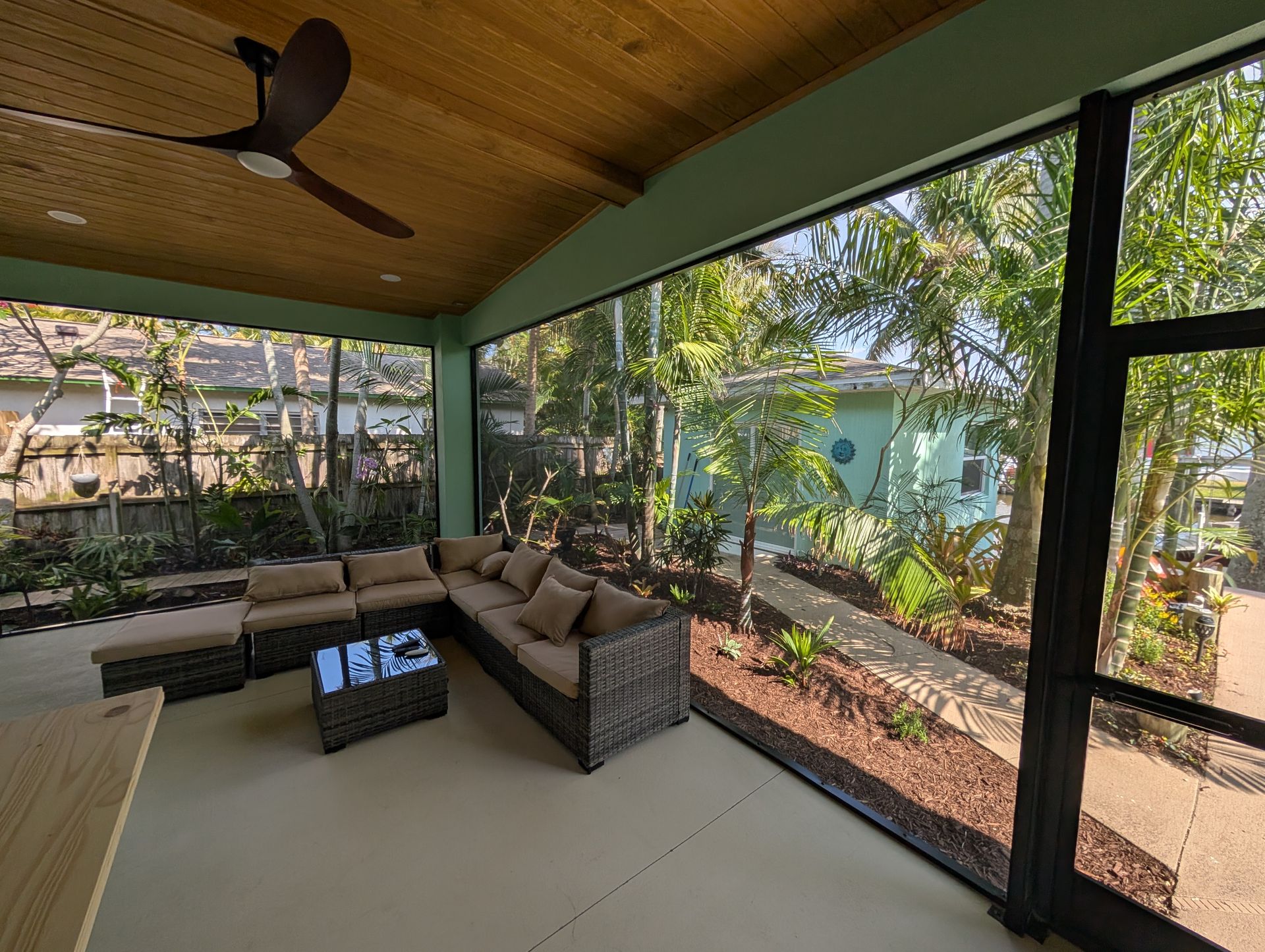 Covered screened patio with wicker sofa, coffee table, and garden view under a wooden ceiling.