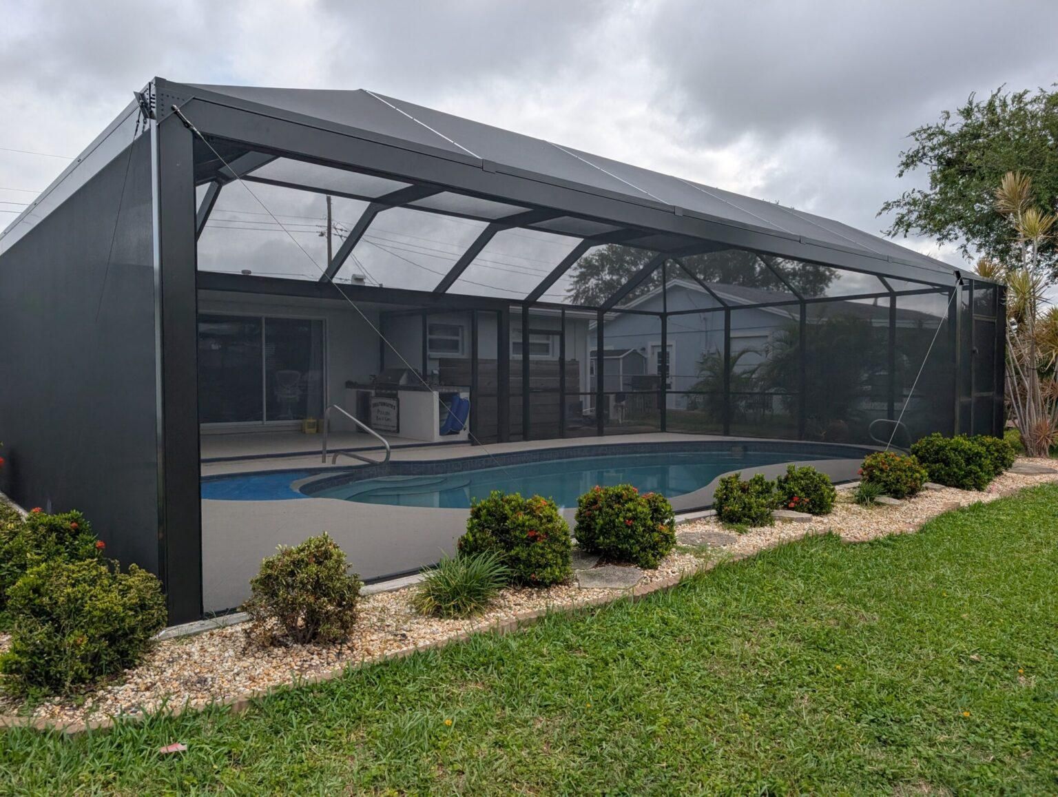 Screened-in backyard pool enclosure with blue pool, dark framing, and grassy landscaping under cloudy sky