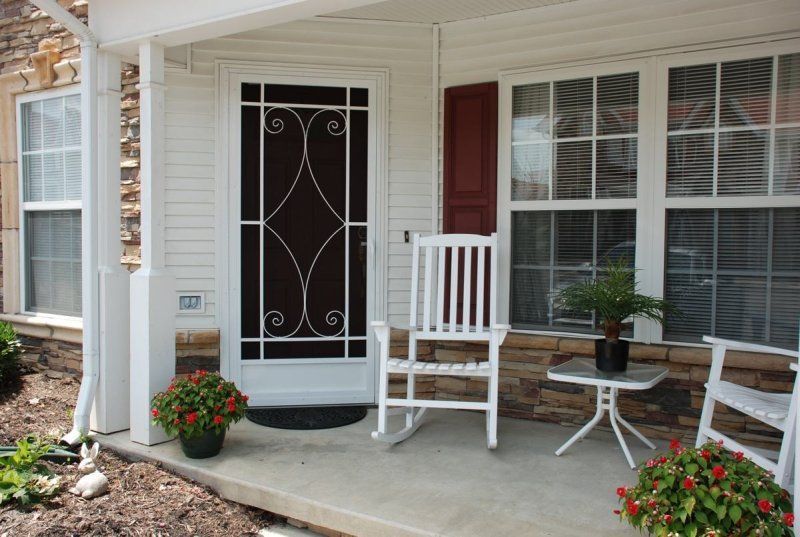 Front porch with white chairs, small table, potted flowers, and a decorative storm door beside large windows.
