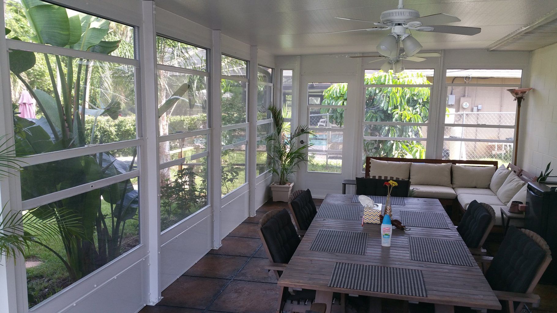Screened porch with long dining table, black chairs, and ceiling fan, overlooking lush greenery.