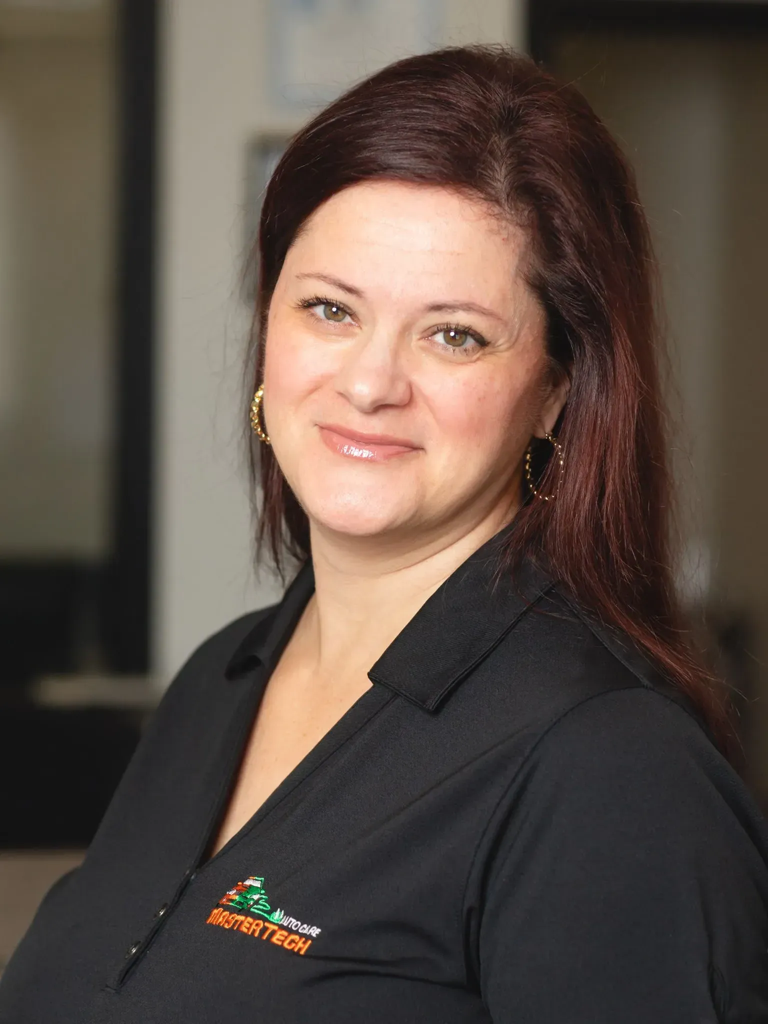 Smiling woman in a black blazer with a logo, standing indoors against a blurred background