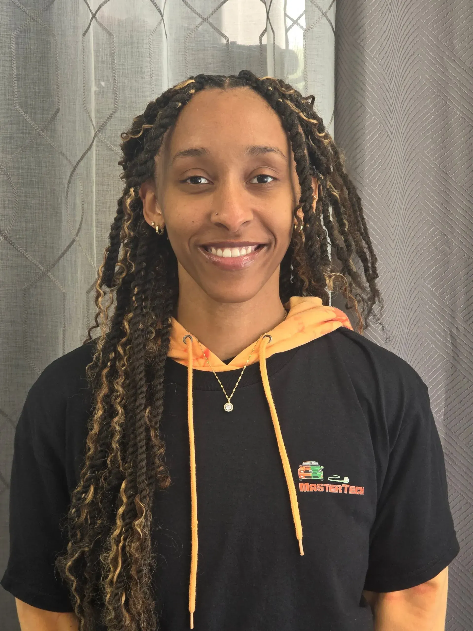 Smiling person in a black shirt with orange hoodie strings, standing before a gray patterned curtain.