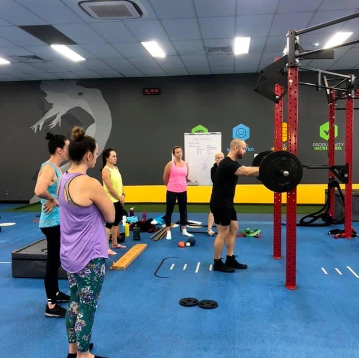 People exercising in a gym, with one person lifting a barbell on a squat rack.