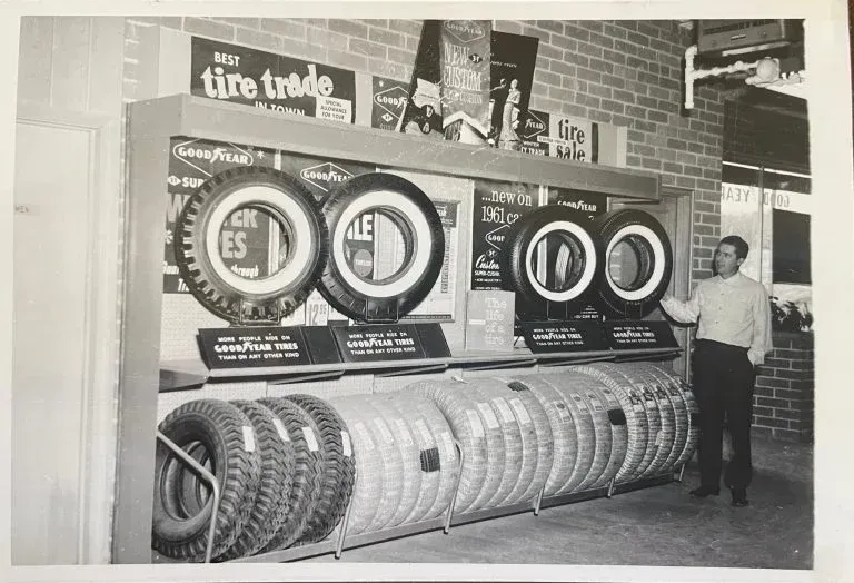 Man standing beside a tire display in a shop, with rows of tires and large circular price signs.
