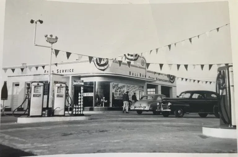 Vintage black-and-white photo of a service station with gas pumps, pennant flags, and parked cars.