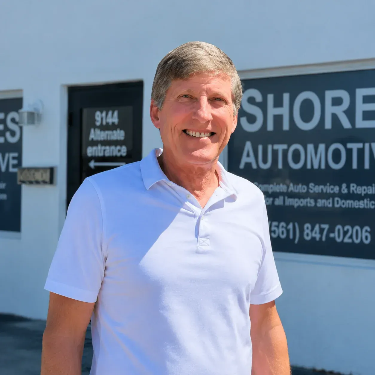 Man in a light blue polo standing outside an automotive shop with signage in the background
