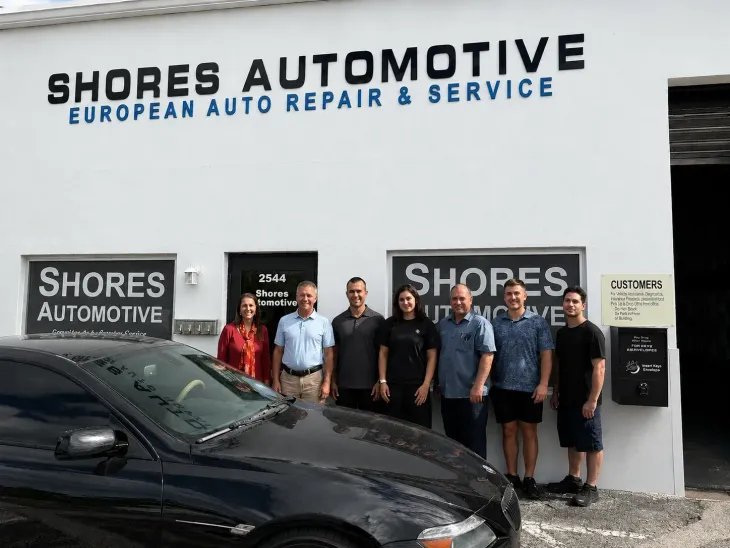 Six people posing outside Shores Automotive in front of a black car and shop signage