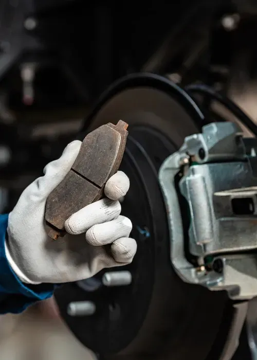 Gloved hand holding a brake pad near a car wheel and brake caliper in a garage