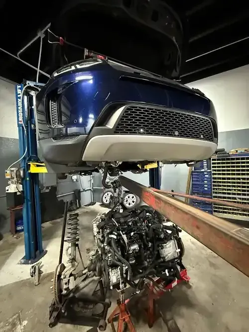 Blue car lifted in a garage, with a damaged engine assembly below it on a work platform