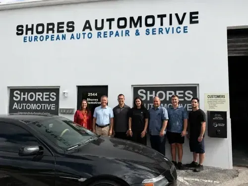 People standing outside Shores Automotive, an auto repair shop, with a black car in front