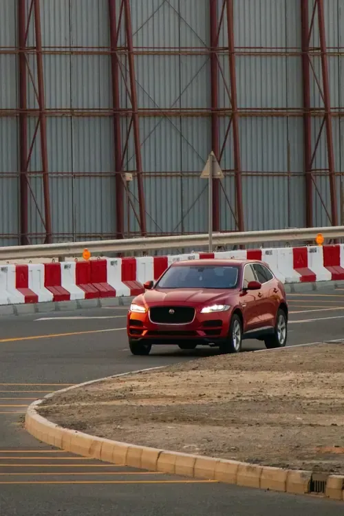 Red SUV driving on a track beside red-and-white barriers and a hangar wall
