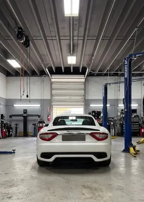 White sports car parked in an auto repair shop with blue vehicle lifts and concrete floor.