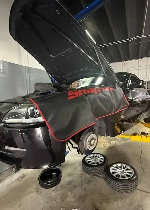 Black car in a repair shop with hood open and Snap-on cover, wheels removed nearby.