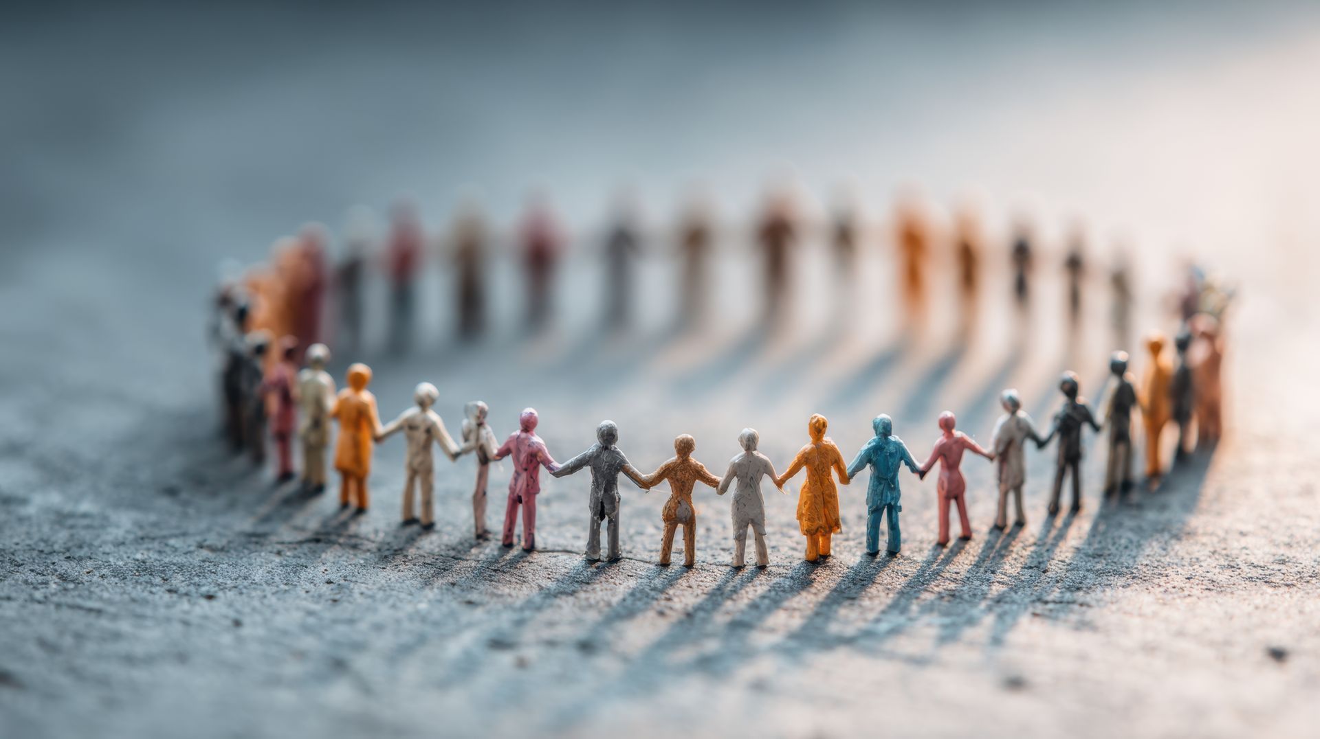 Small, multicolored toy figures hold hands in a large, blurred circle on a textured, gray surface.