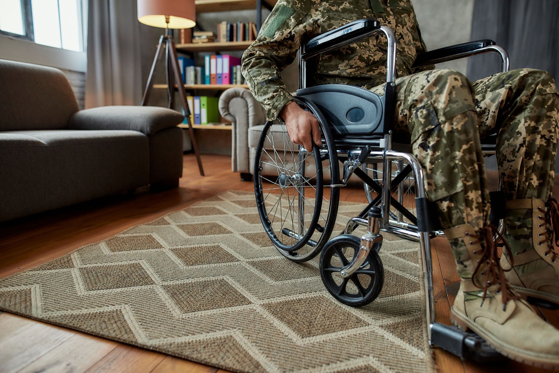 A person in camouflage uniform sitting in a wheelchair on a patterned rug in a room with a sofa and bookshelf.