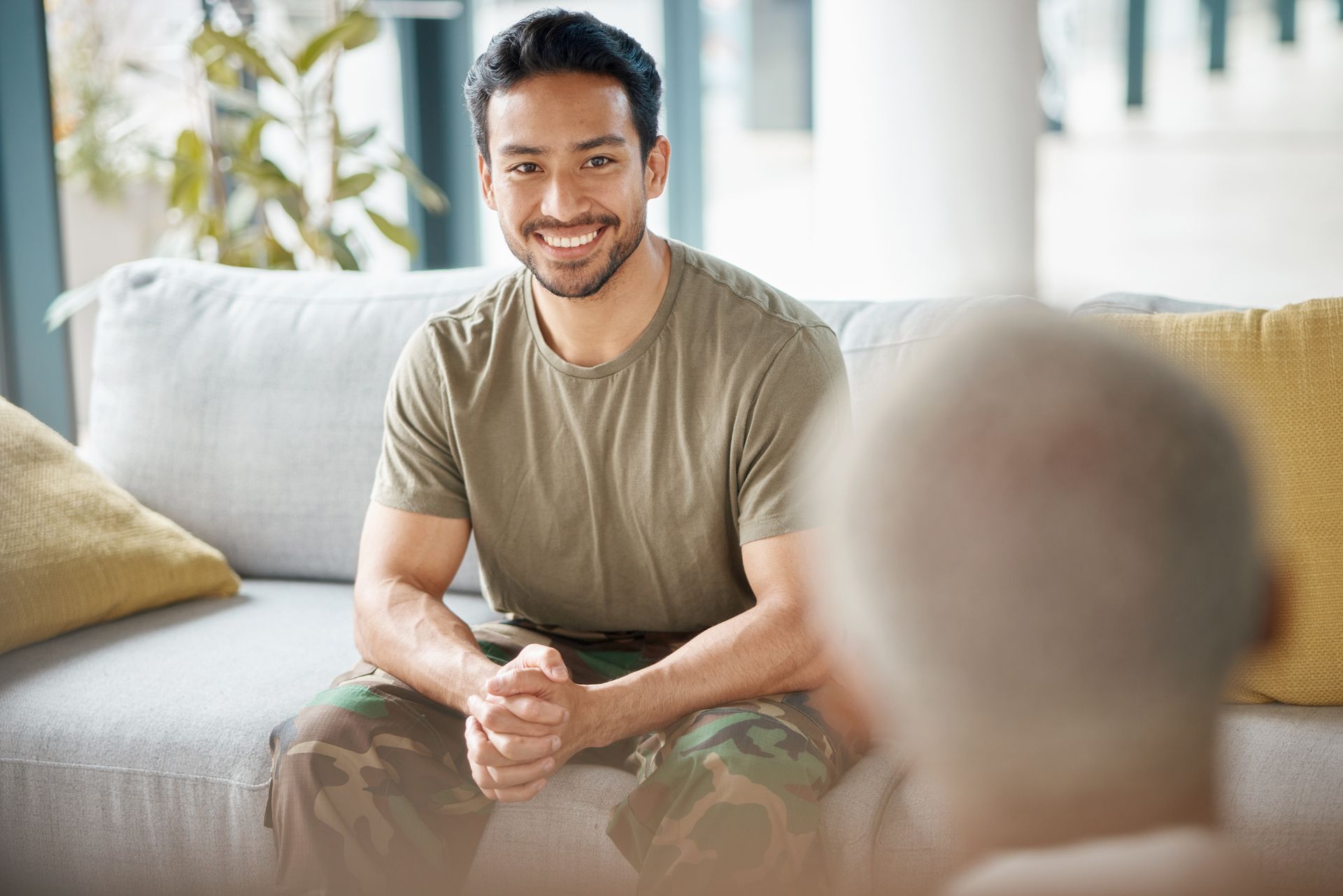 A person in a camouflage uniform sits on a sofa, smiling at a person whose back is to the camera.