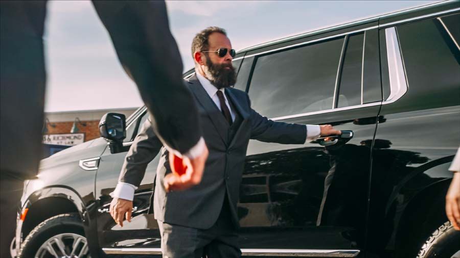 Man in a dark suit opening the door of a black SUV while another person stands nearby.