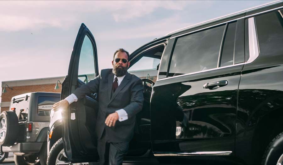 Man in sunglasses stepping from a black SUV with the driver’s door open, wearing a dark suit.