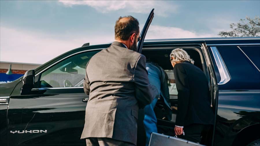 Two men in suits stand by an open black SUV, one helping the other get in.
